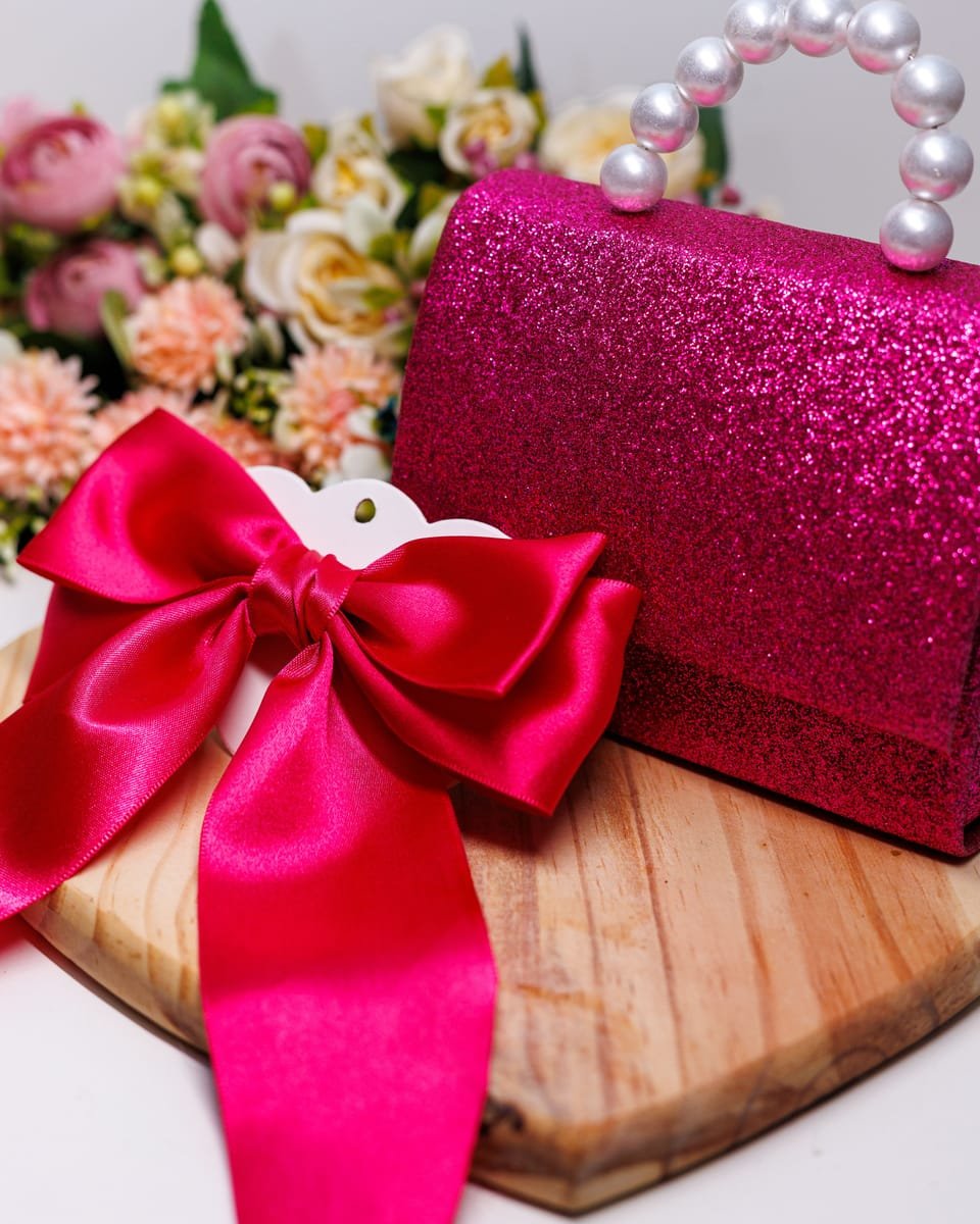 Laço de cetim pink ao lado de uma bolsinha de glitter pink com alça de pérolas